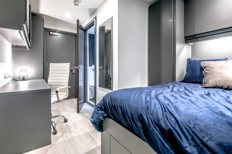 A large studio bedroom featuring a double bed, study desk, wardrobe, and ample storage space