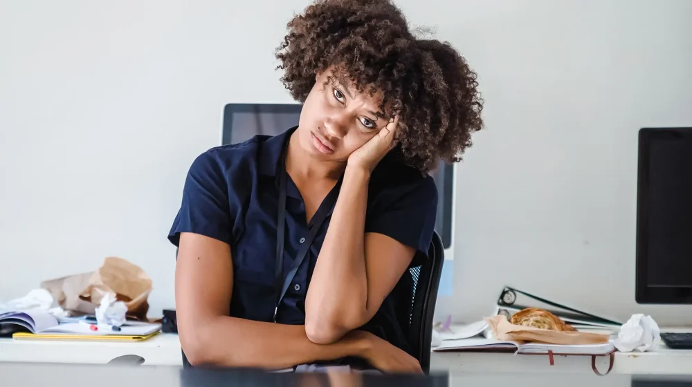 Exhausted student resting her head on her hand at a cluttered desk with a computer and scattered papers